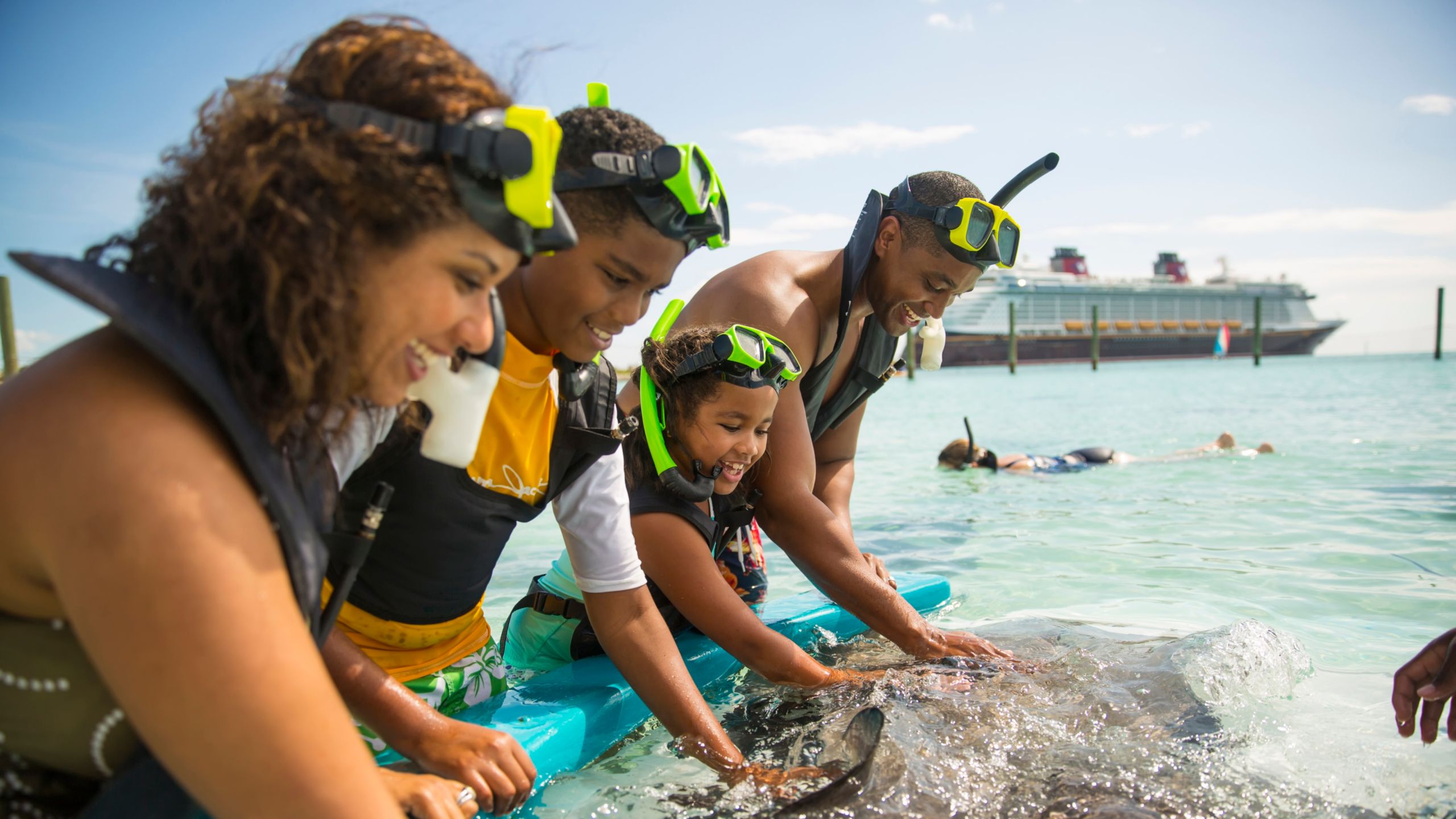 A family of 4 wearing snorkeling gear interacting with a stingray at Disney Castaway Cay, with a Disney Cruise Line ship in the background