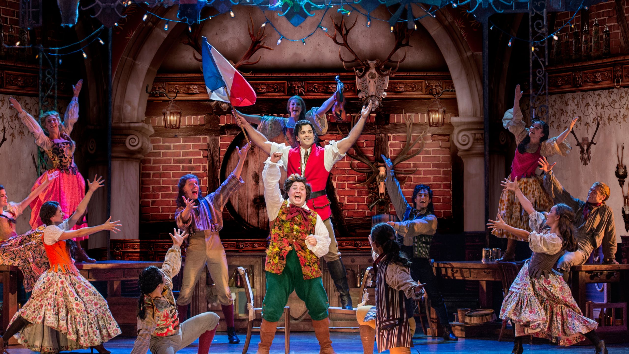 Gaston segurando uma bandeira da França com os braços levantados ao lado de LeFou no palco de Beauty and the Beast