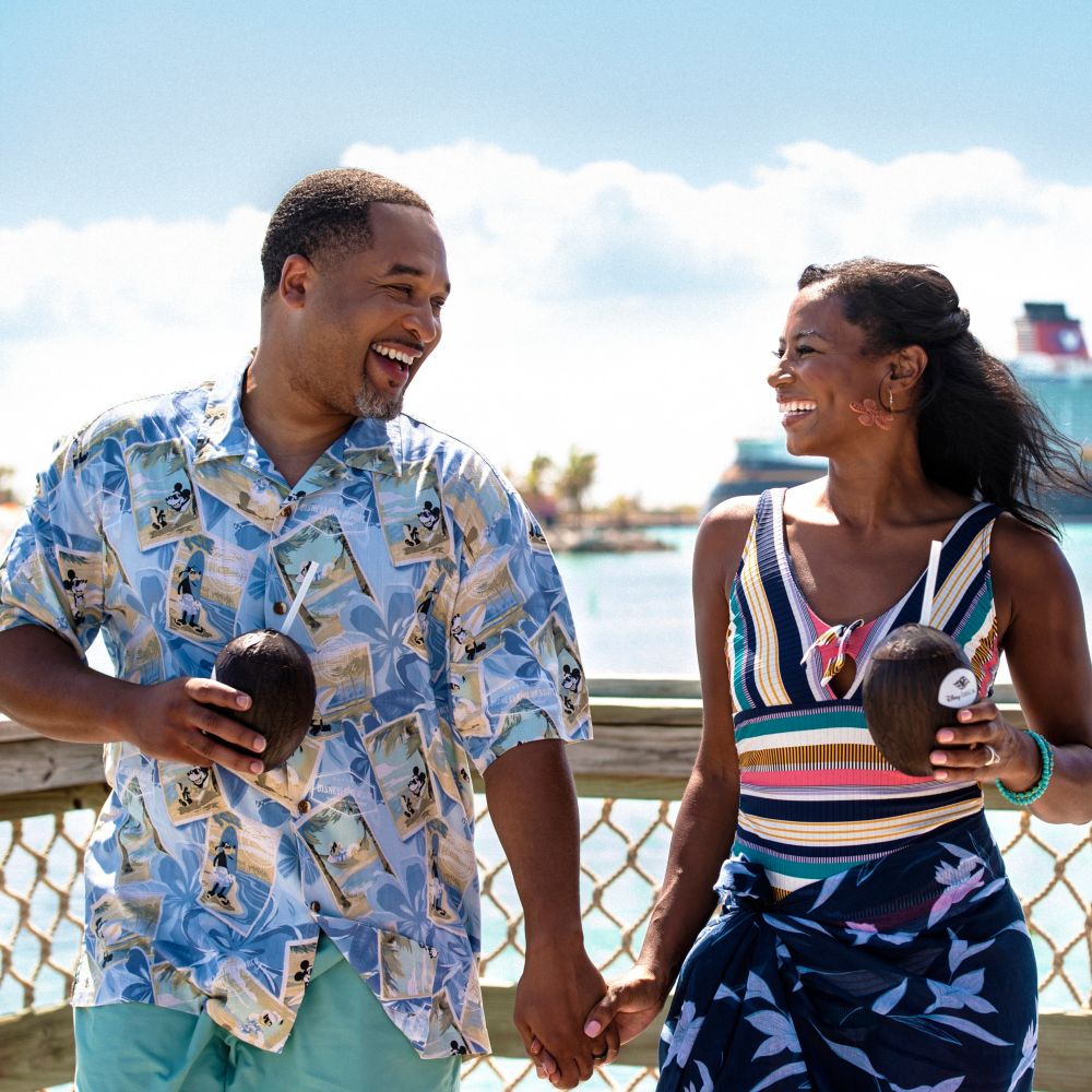 A man and a woman holding hands and drinking from coconuts with a Disney Cruise Line ship behind them