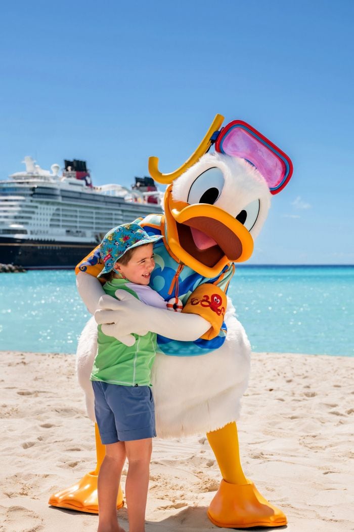 Donald Duck hugging a young Guest on a beach with a docked Disney Cruise Line ship