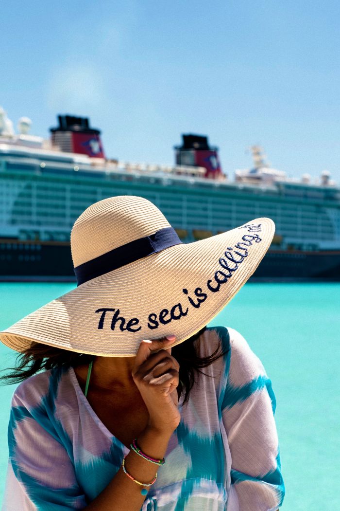 Uma mulher na praia, diante de um navio de cruzeiro da Disney, abaixando a borda de seu chapéu de sol e revelando a frase: "O mar me chama" em inglês