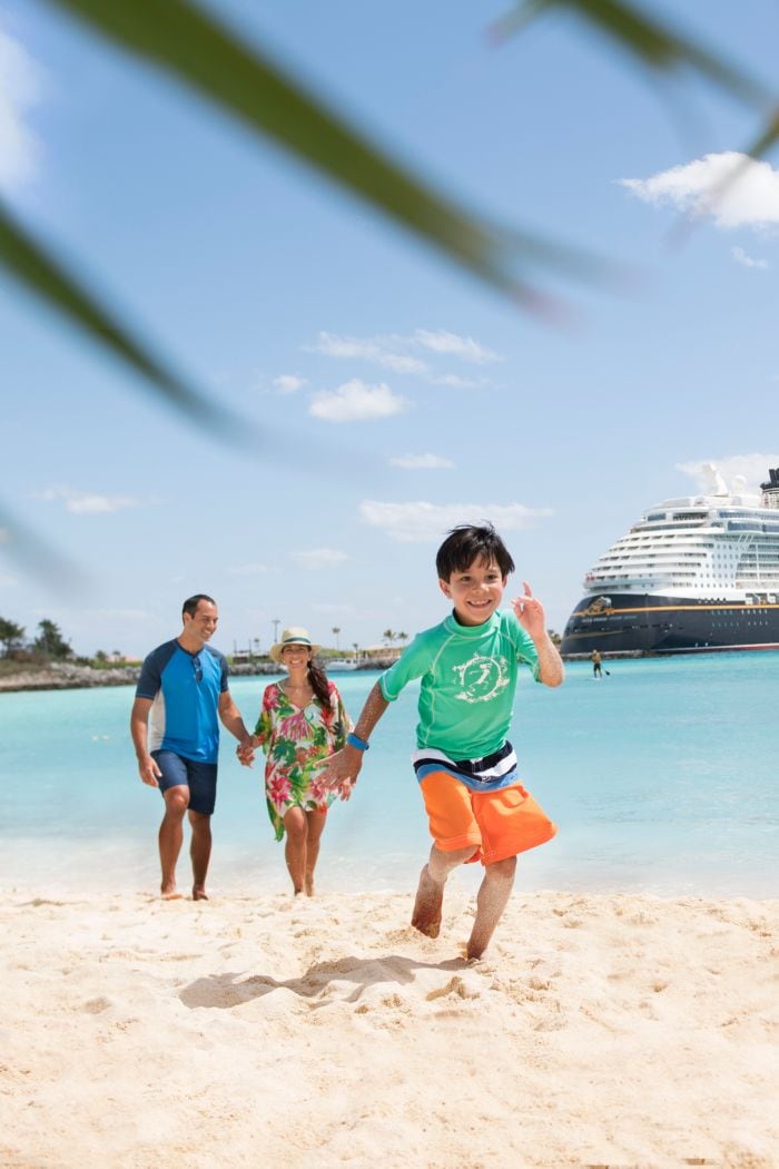 A child runs on the beach in front of his parents with the Disney Fantasy cruise ship docked behind them
