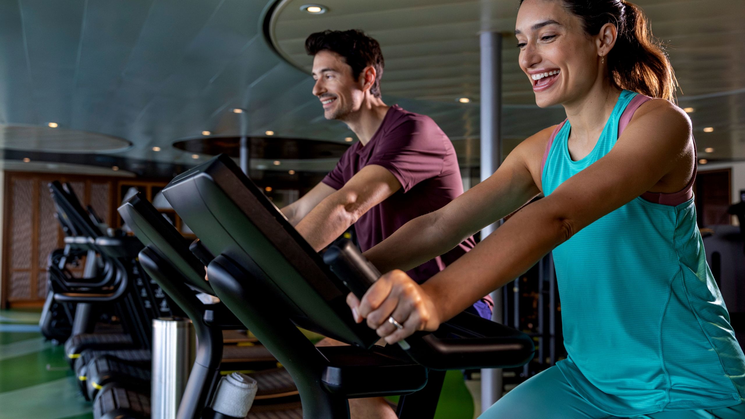 A smiling man and woman ride stationary bicycles in a gym  