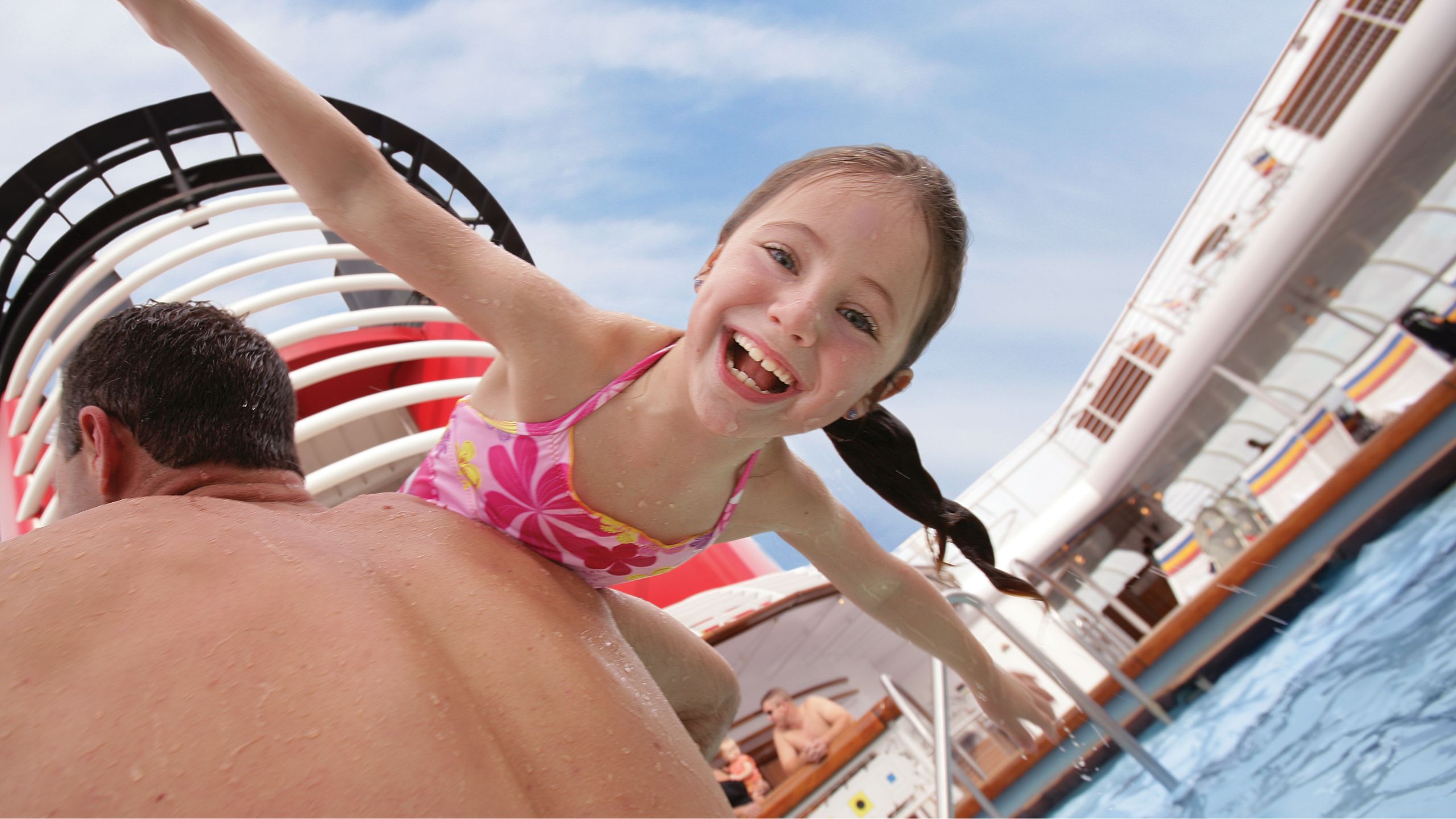 A smiling girl hangs over her father’s shoulder in a pool on the deck of a Disney Cruise Line ship 