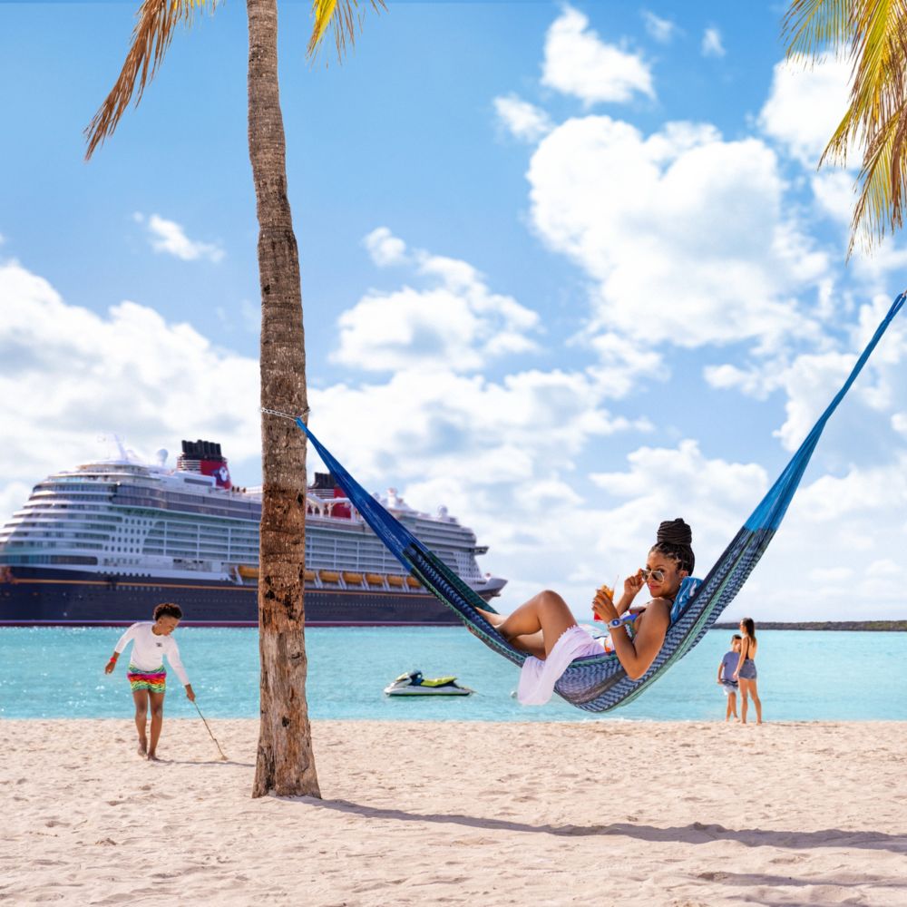A woman lounges in a hammock on the beach in front of the Disney Wish cruise ship