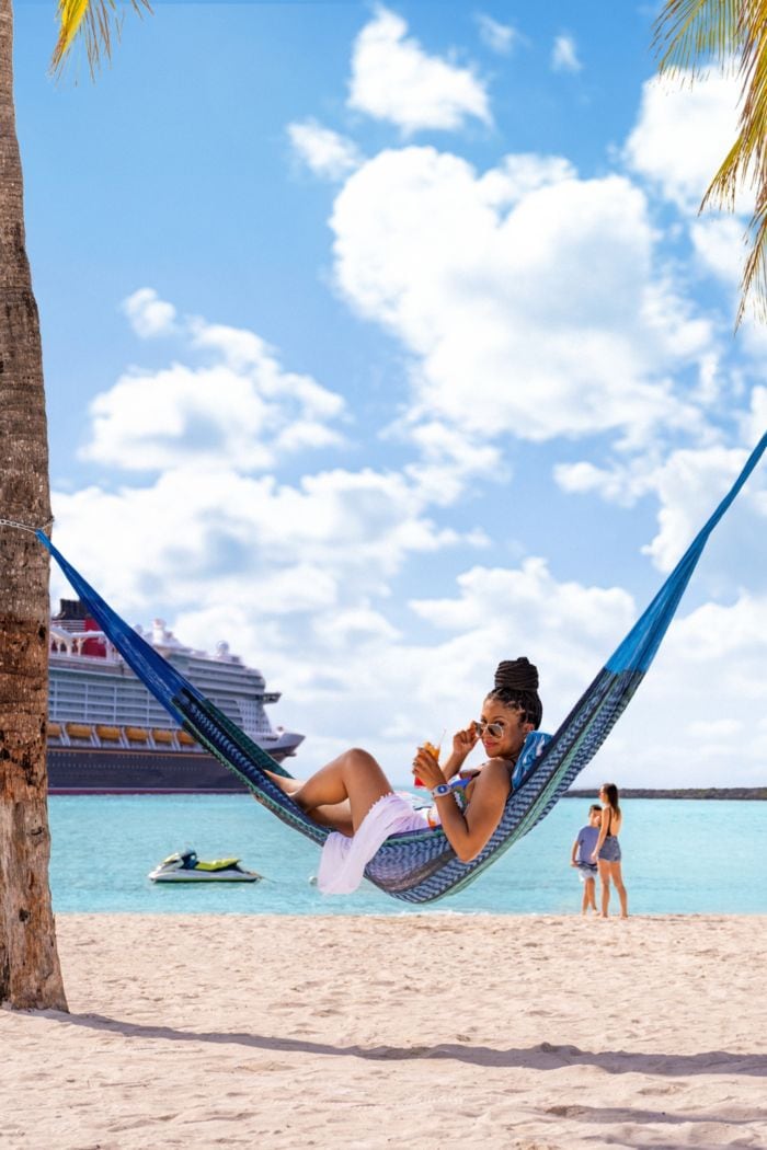 A woman lounges in a hammock on the beach in front of the Disney Wish cruise ship