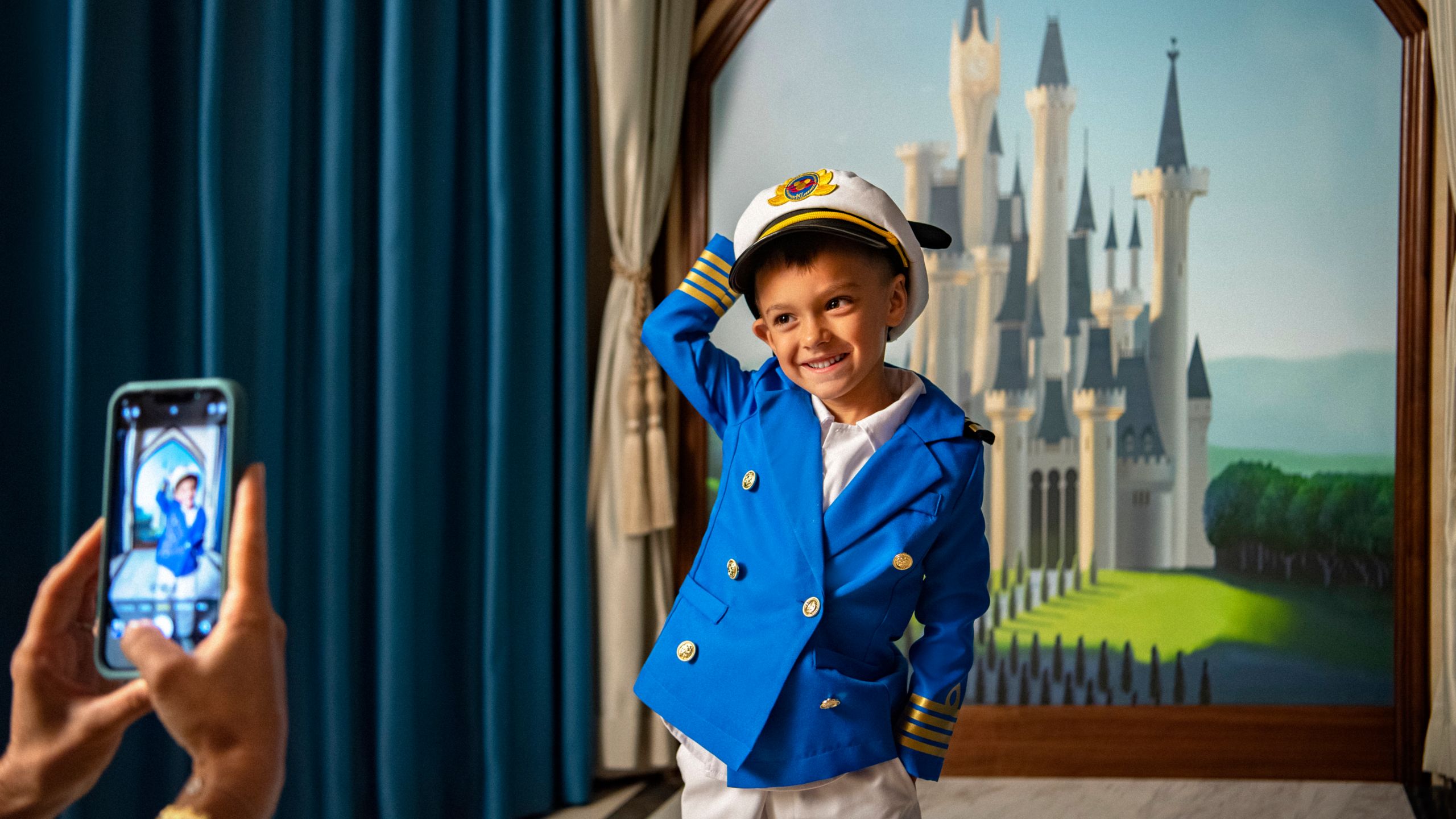 A young boy dressed in a Captain's uniform, having his picture taken in front of a painting of a castle