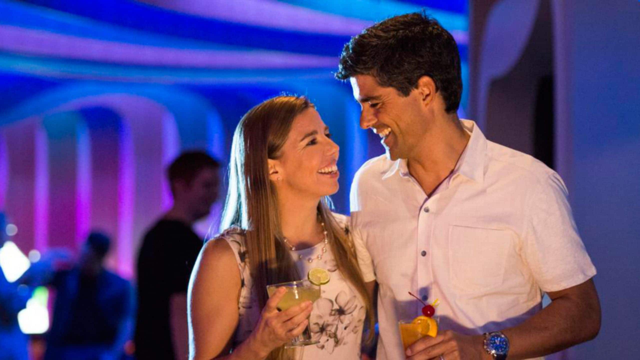 Um casal de pé lado a lado segurando bebidas e se olhando nos olhos