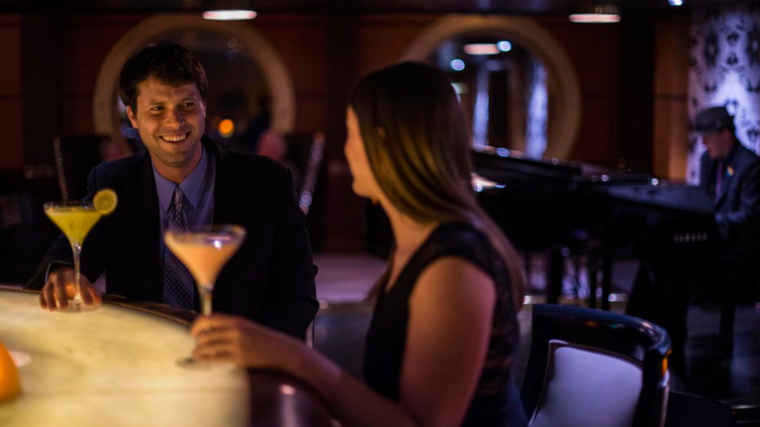 Um casal sentado no bar de um lounge com suas bebidas enquanto um pianista toca ao fundo