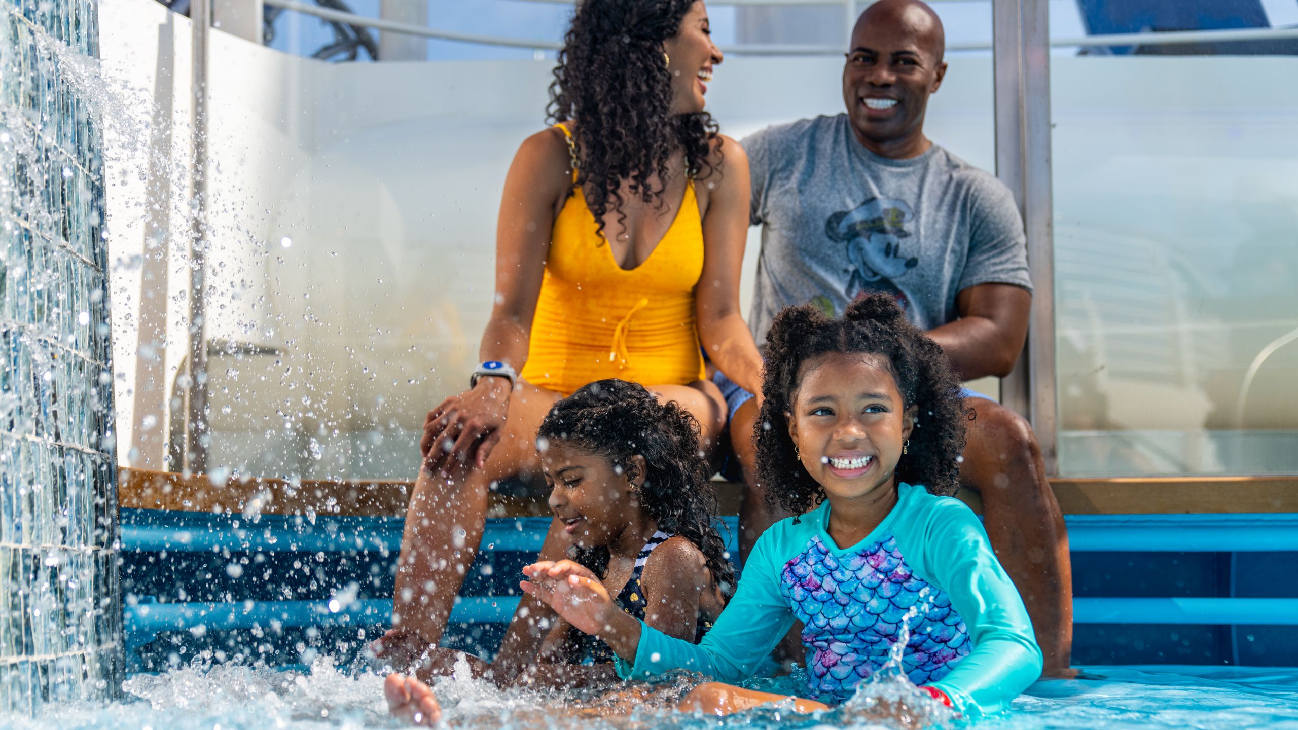 Two young girls sitting with their parents and splashing in shallow water on a Disney cruise ship
