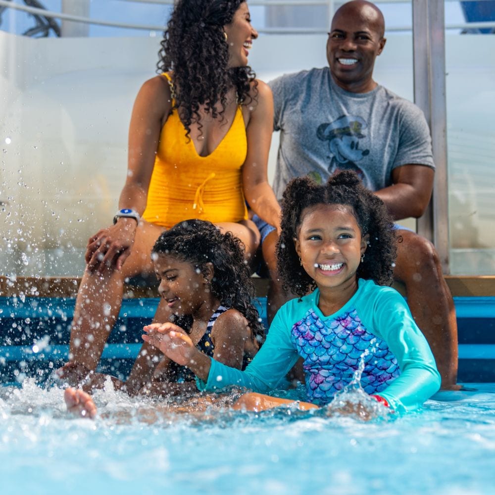 Two young girls sitting with their parents and splashing in shallow water on a Disney cruise ship