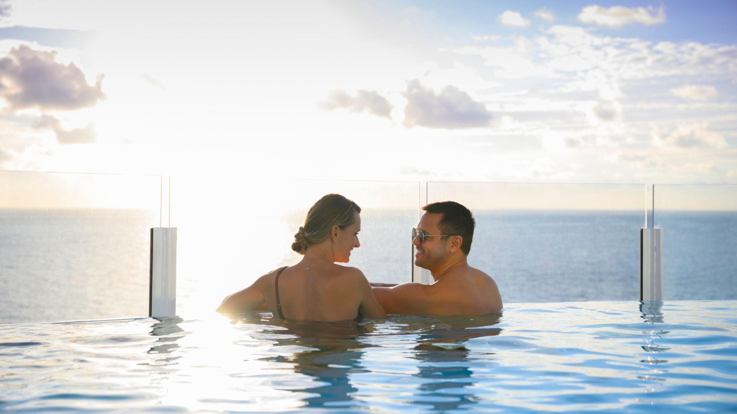 Un homme et une femme debout dans une piscine à débordement face à l’océan au coucher du soleil