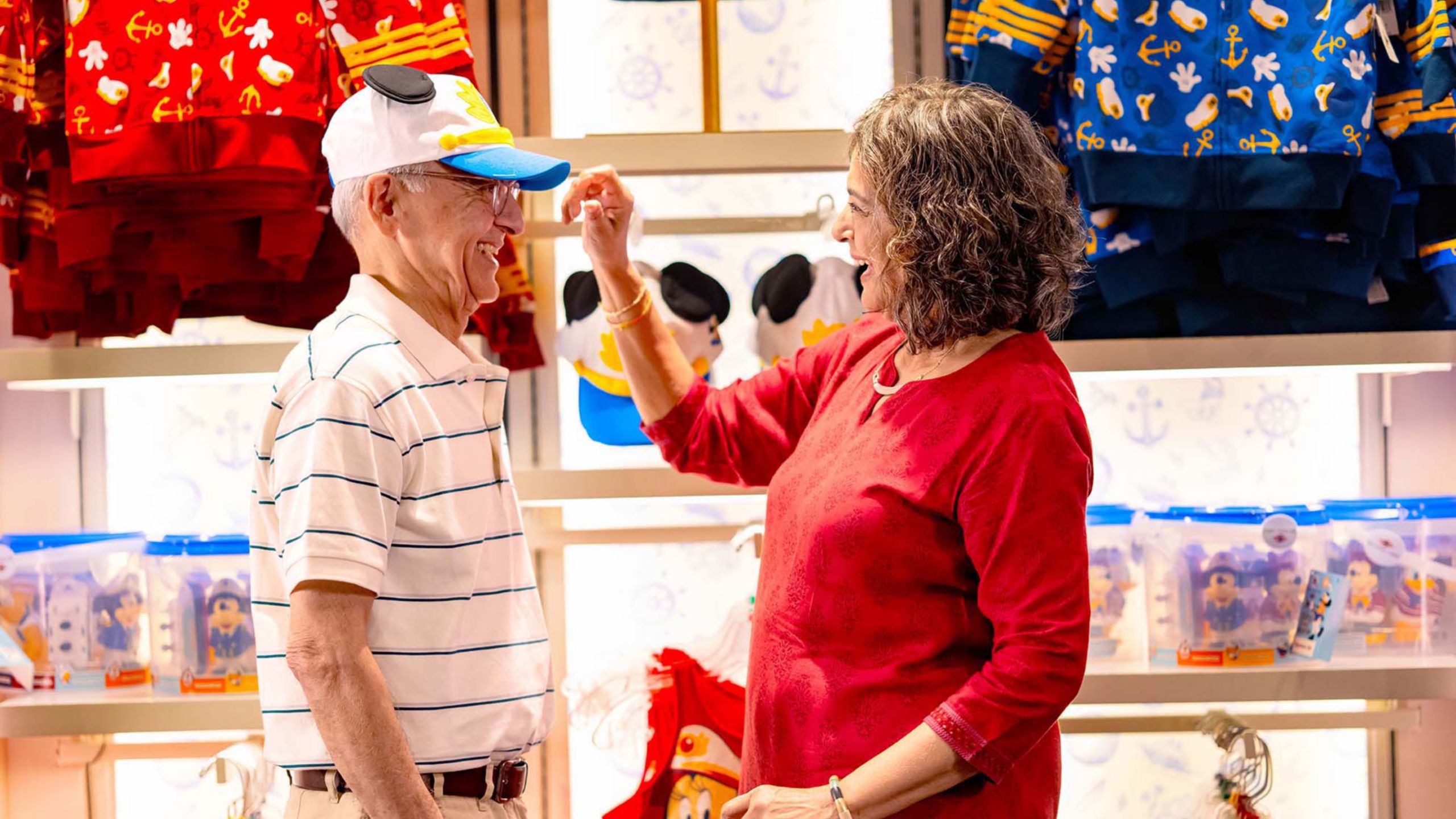 A woman puts a hat with Mickey Mouse ears on a man’s head while standing inside a giftshop on board the Disney Adventure