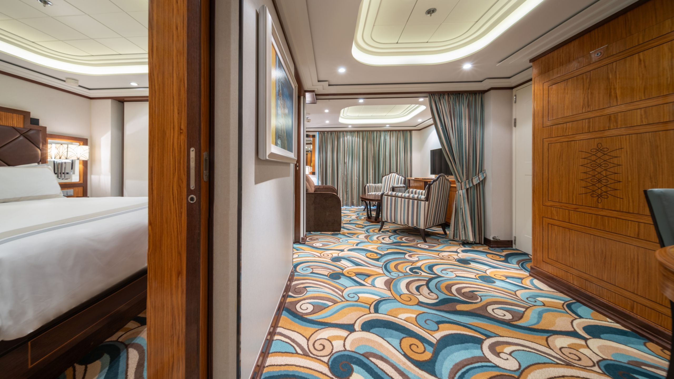 A bedroom with a bed, a nightstand, a sofa, 2 chairs and a TV in a Disney Dream Concierge 1 Bedroom Suite with Verandah