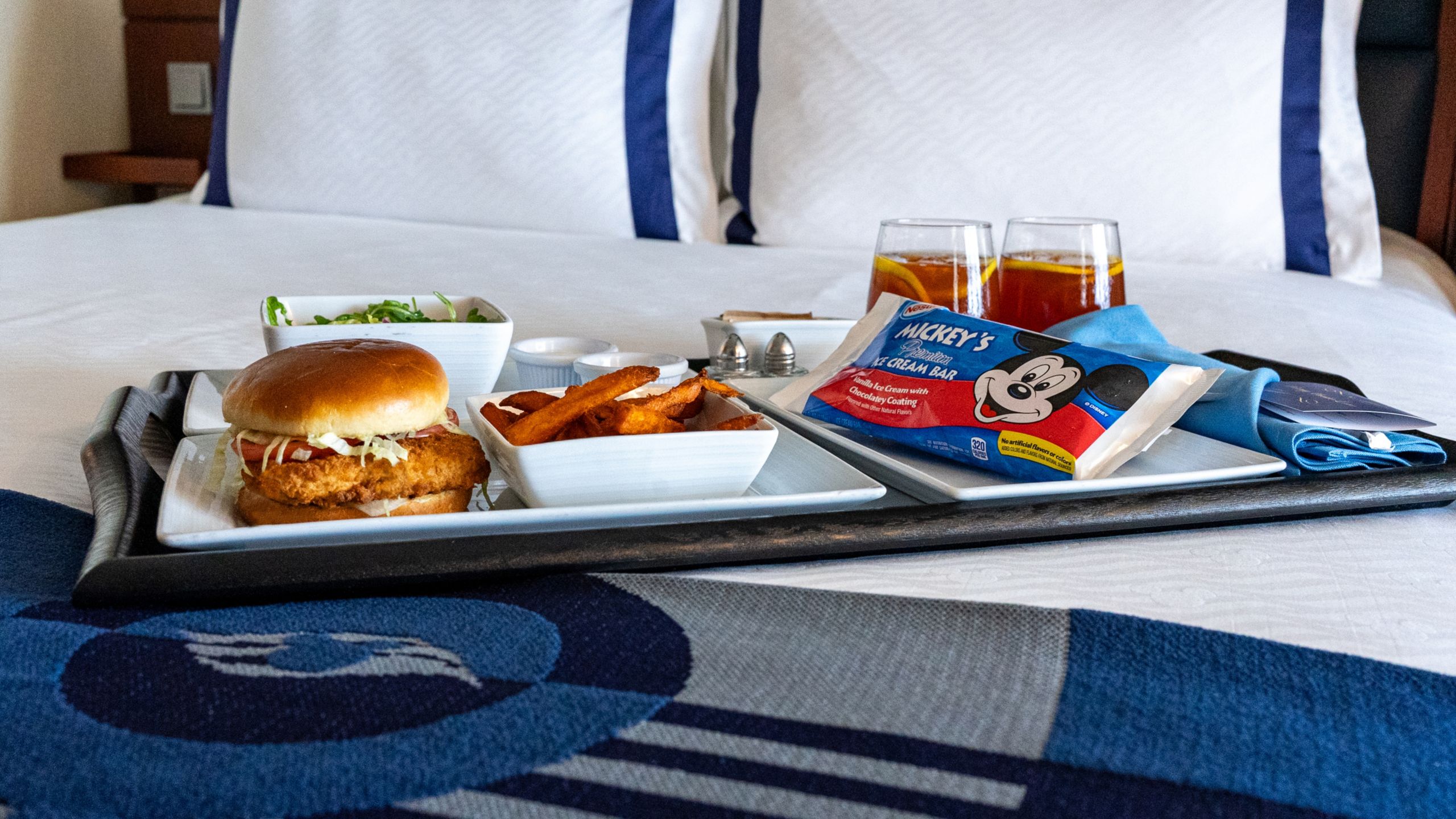 A tray with a hamburger, a plate of chips, a salad, 2 beverages and ice cream on the bed of a Disney Fantasy Standard Inside Stateroom