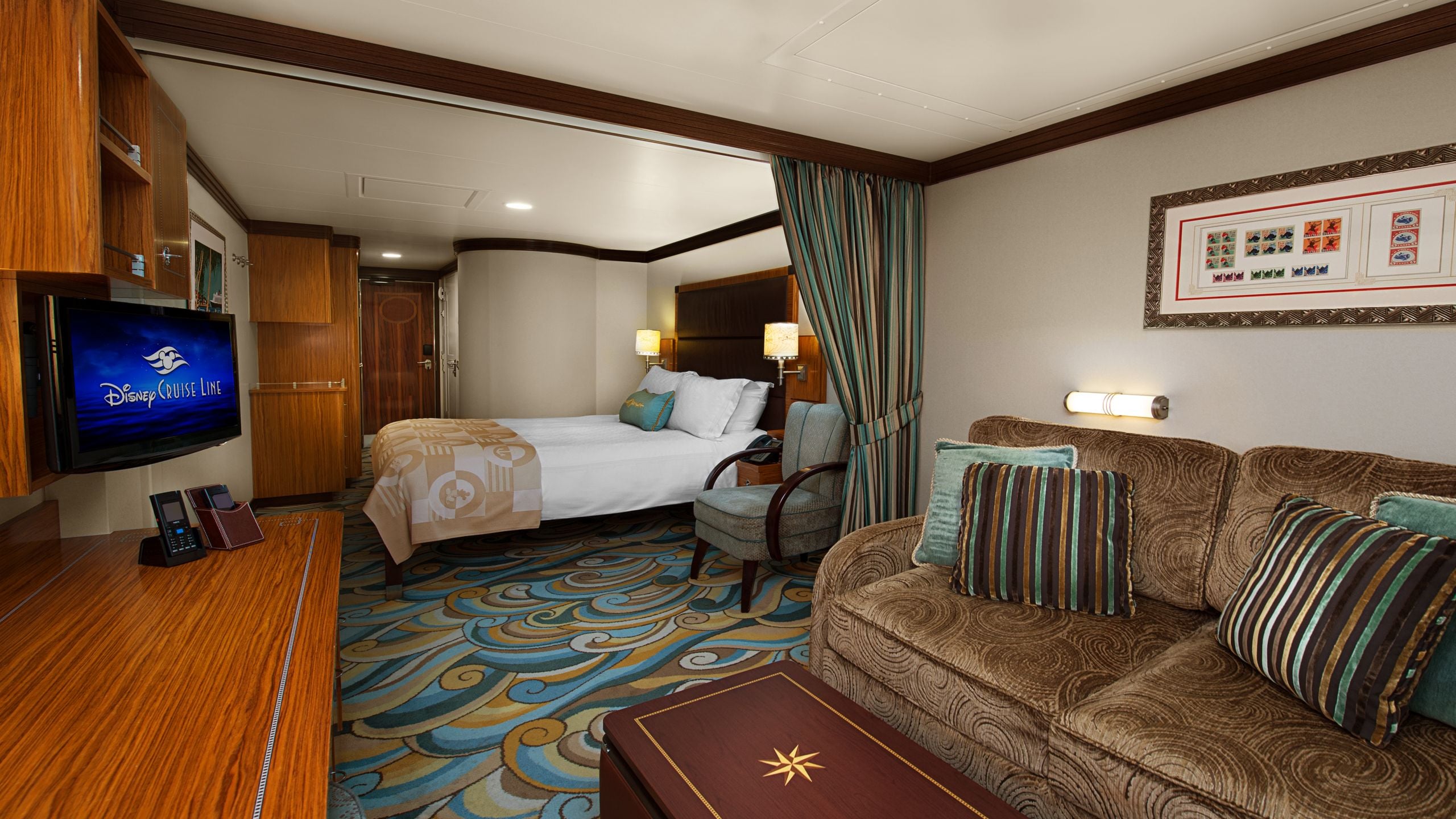 A stateroom with a queen bed, 2 lamps, 2 nightstands, a sofa, a desk and a TV