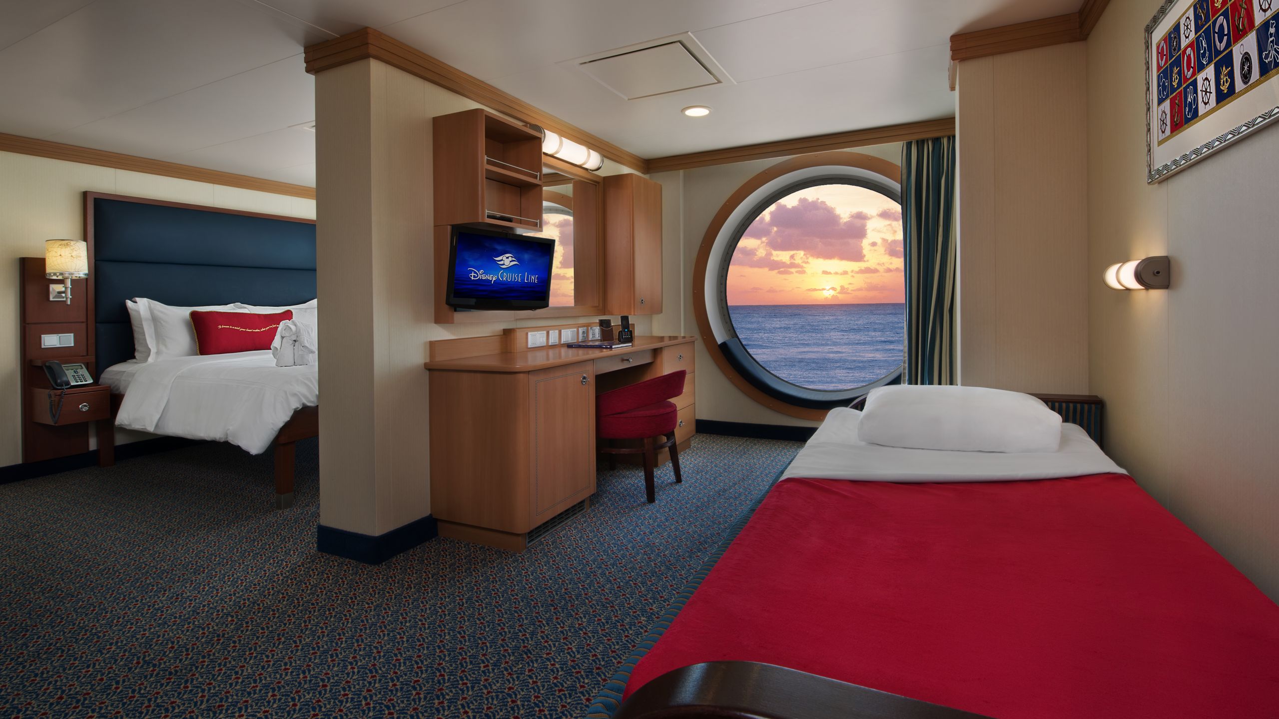 A stateroom with a queen bed and a sofa bed separated by a built in wooden desk and a TV next to a large porthole window