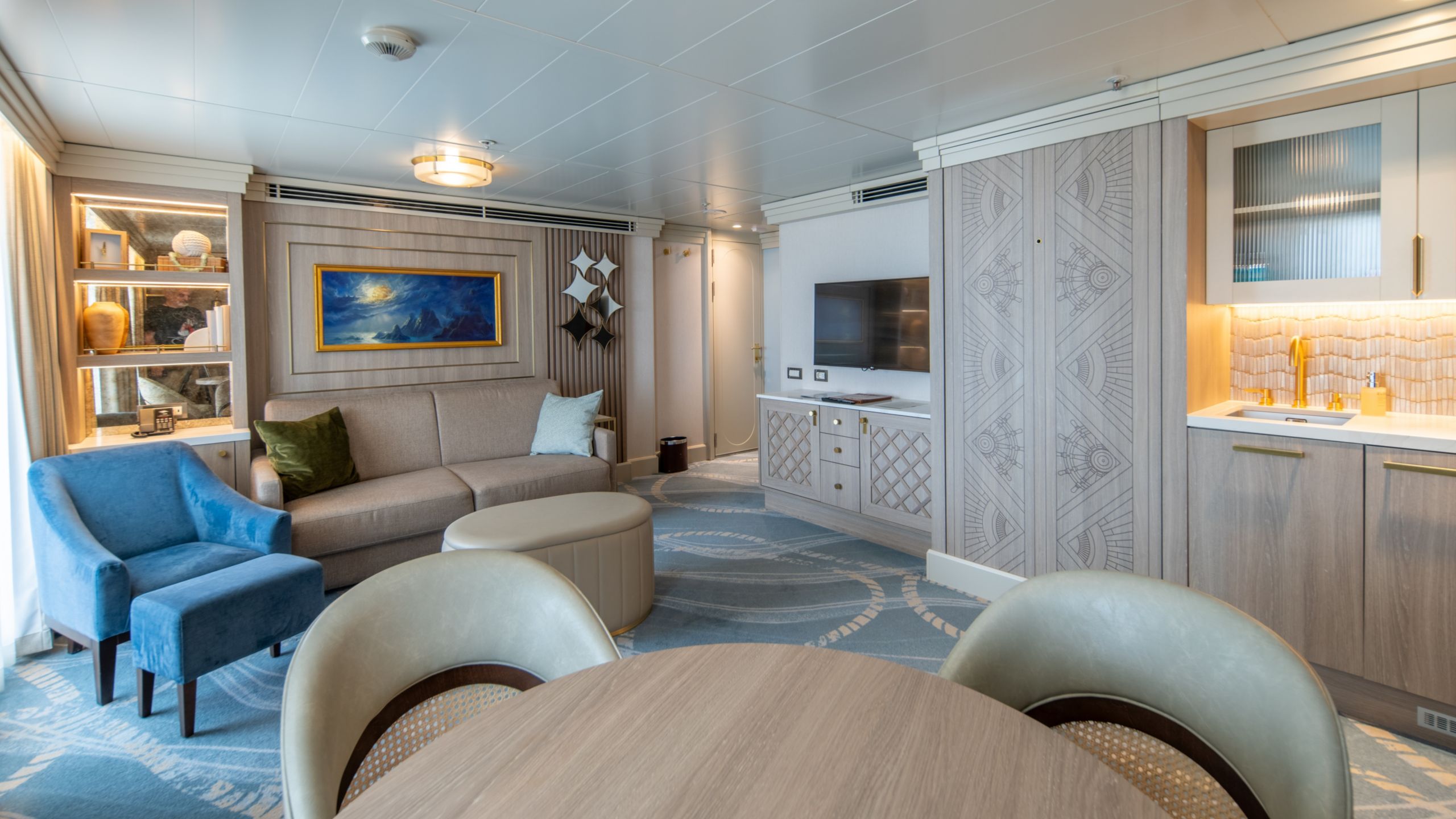 A living area with a wall bed, sofa bed, dining table, TV, wet bar and 3 chairs in a Disney Magic Concierge 1 Bedroom Suite with Verandah