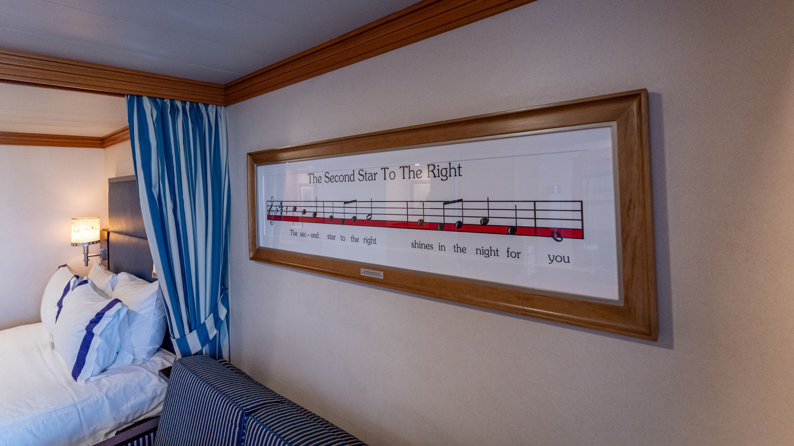 A Disney Magic Deluxe Oceanview with Verandah Stateroom with a bed and artwork above a sofa featuring sheet music from The Second Star to the Right