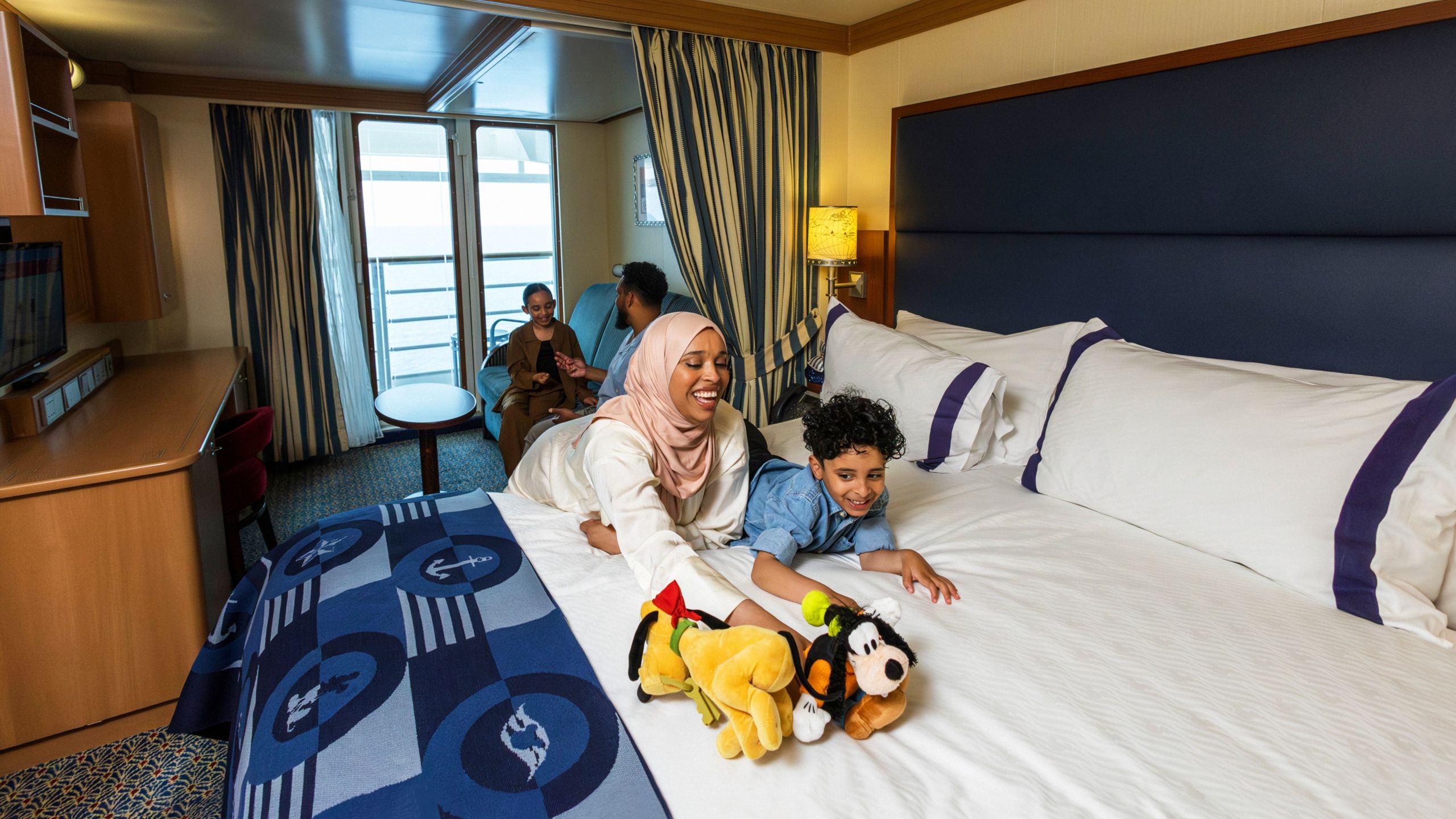 A family relaxing in a Disney Cruise Line stateroom with a bed, sofa, table, desk, TV, chair and a verandah facing the ocean