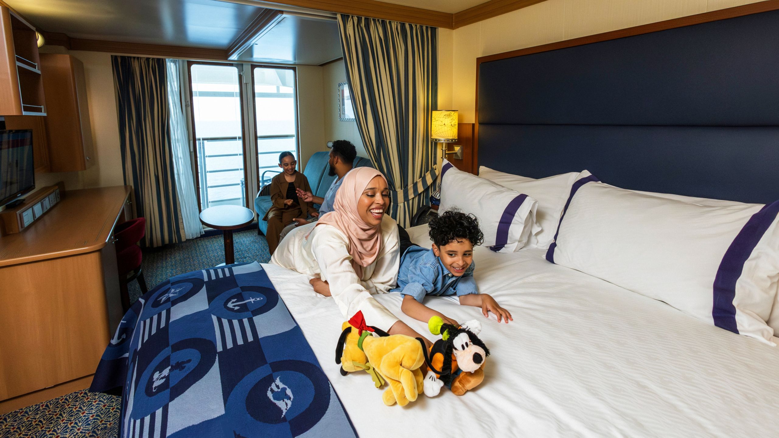 A family relaxing in a Disney Cruise Line stateroom with a bed, sofa, table, desk, TV, chair and a verandah facing the ocean