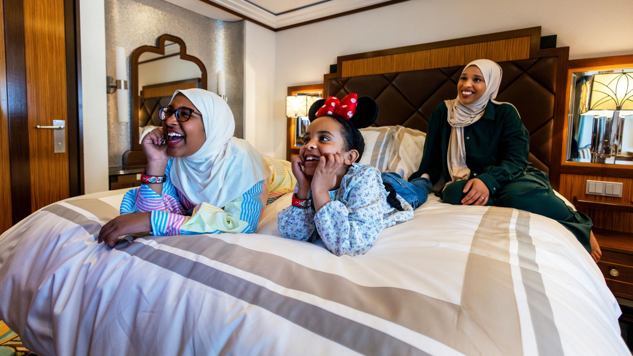 Two girls and their mother smiling while relaxing on a bed in a Disney Cruise Line stateroom