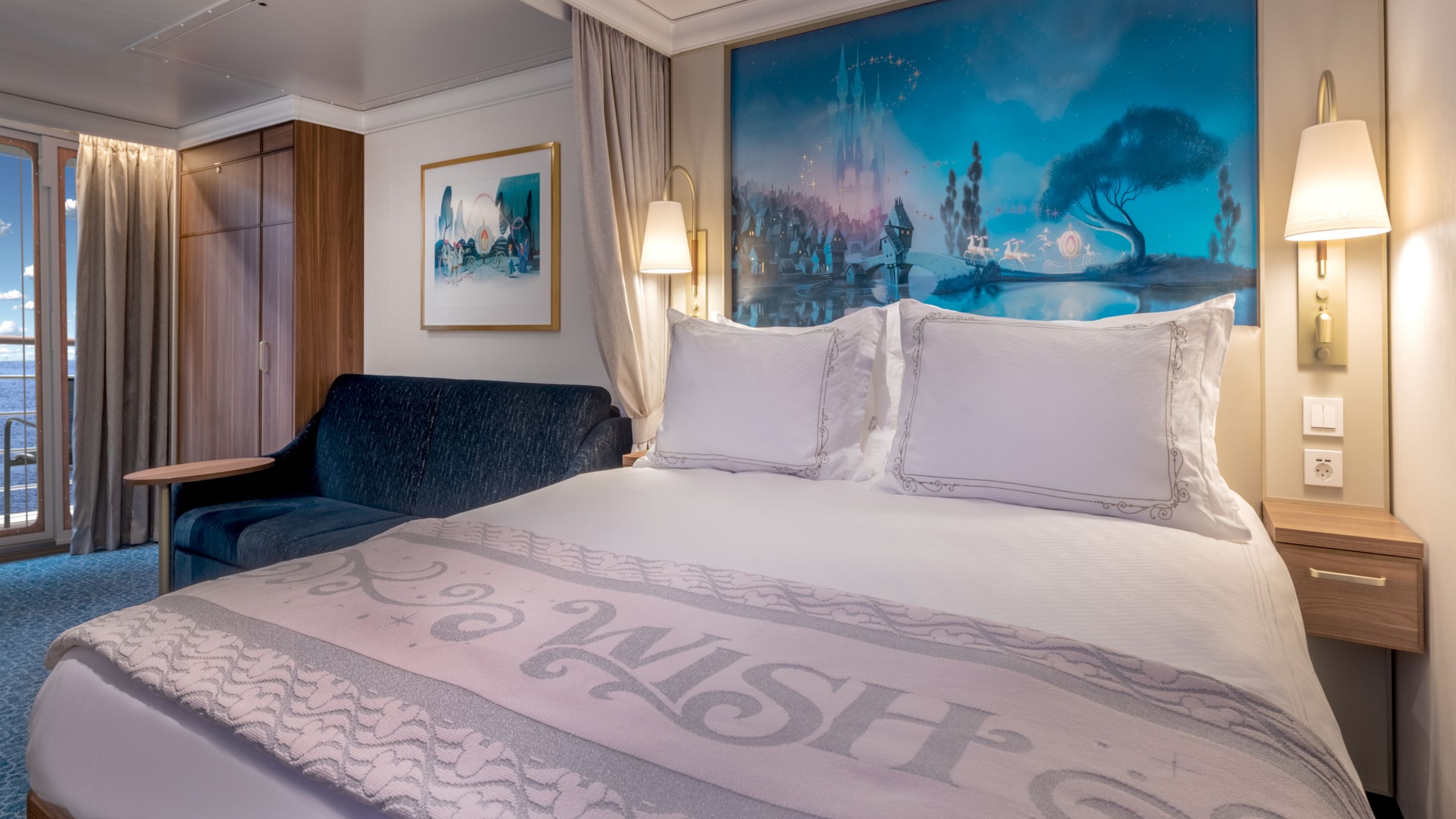 A Disney Wish Verandah Stateroom with a sofa, artwork inspired by Cinderella, table, verandah, 2 nightstands and a bed with luxury bedding