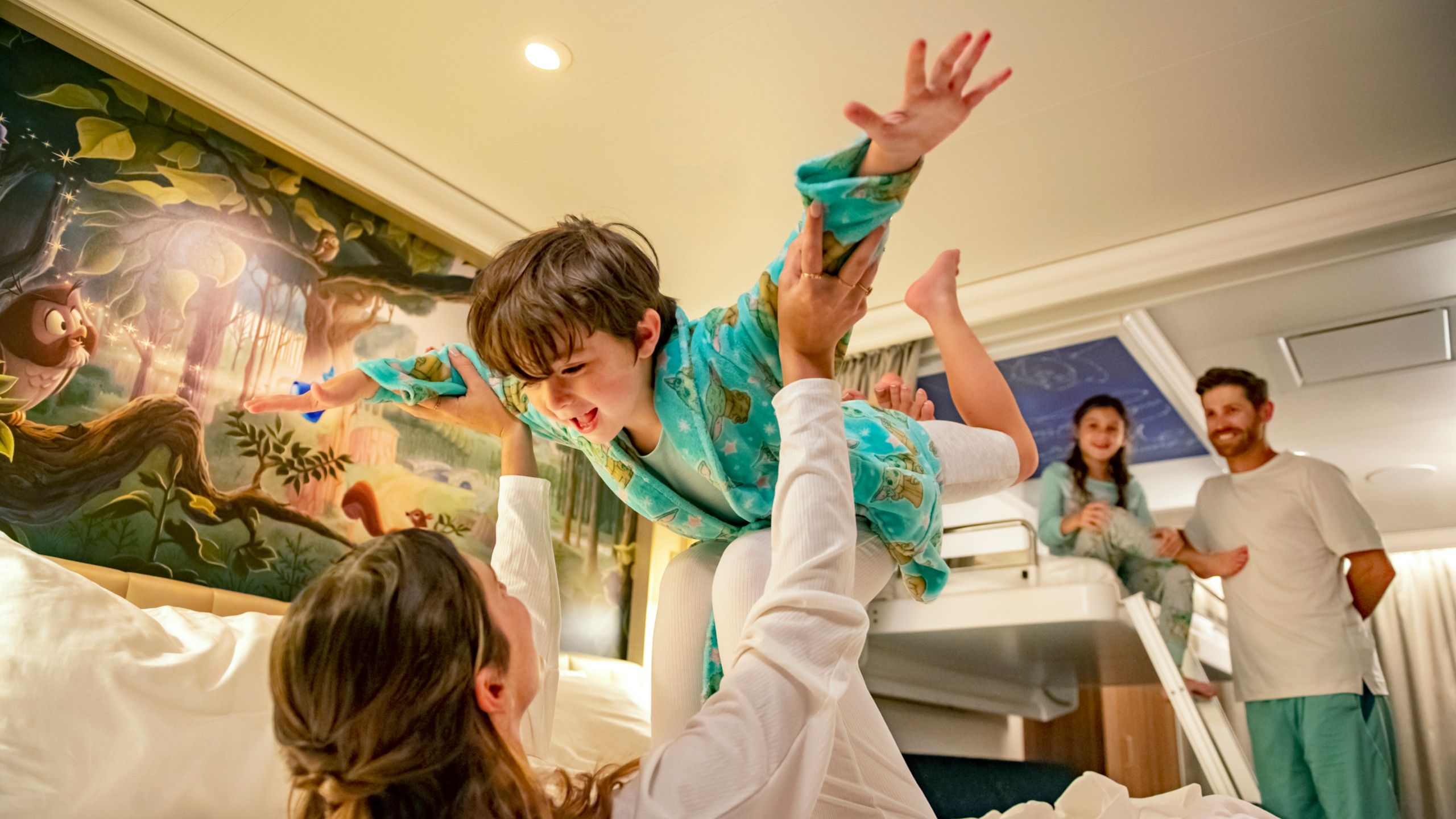 A mother playing with her son on a bed while their father and sister watch in a Disney Cruise Line stateroom with artwork inspired by Sleeping Beauty