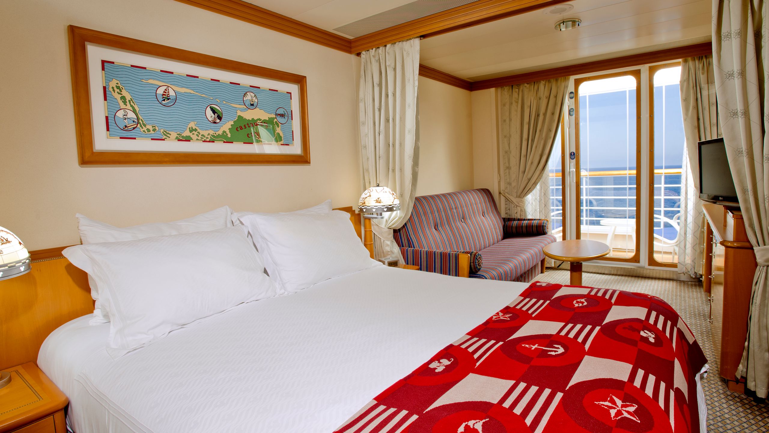 Uma cabine com varanda do Disney Wonder, com cama, mesa, TV, sofá, escrivaninha, 2 mesas de cabeceira e uma varanda de frente para o mar