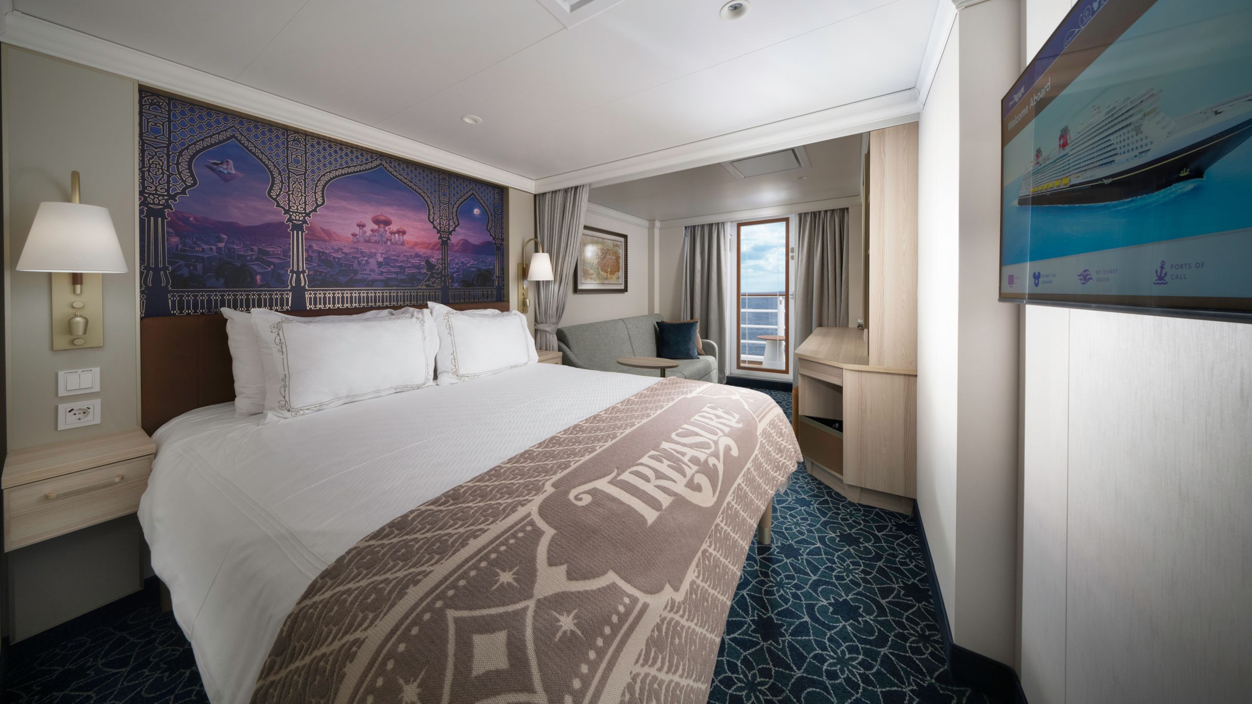 Un camarote del Disney Treasure con una cama, sofá, escritorio, balcón, dos mesas de noche y obras de arte inspiradas en Aladdin