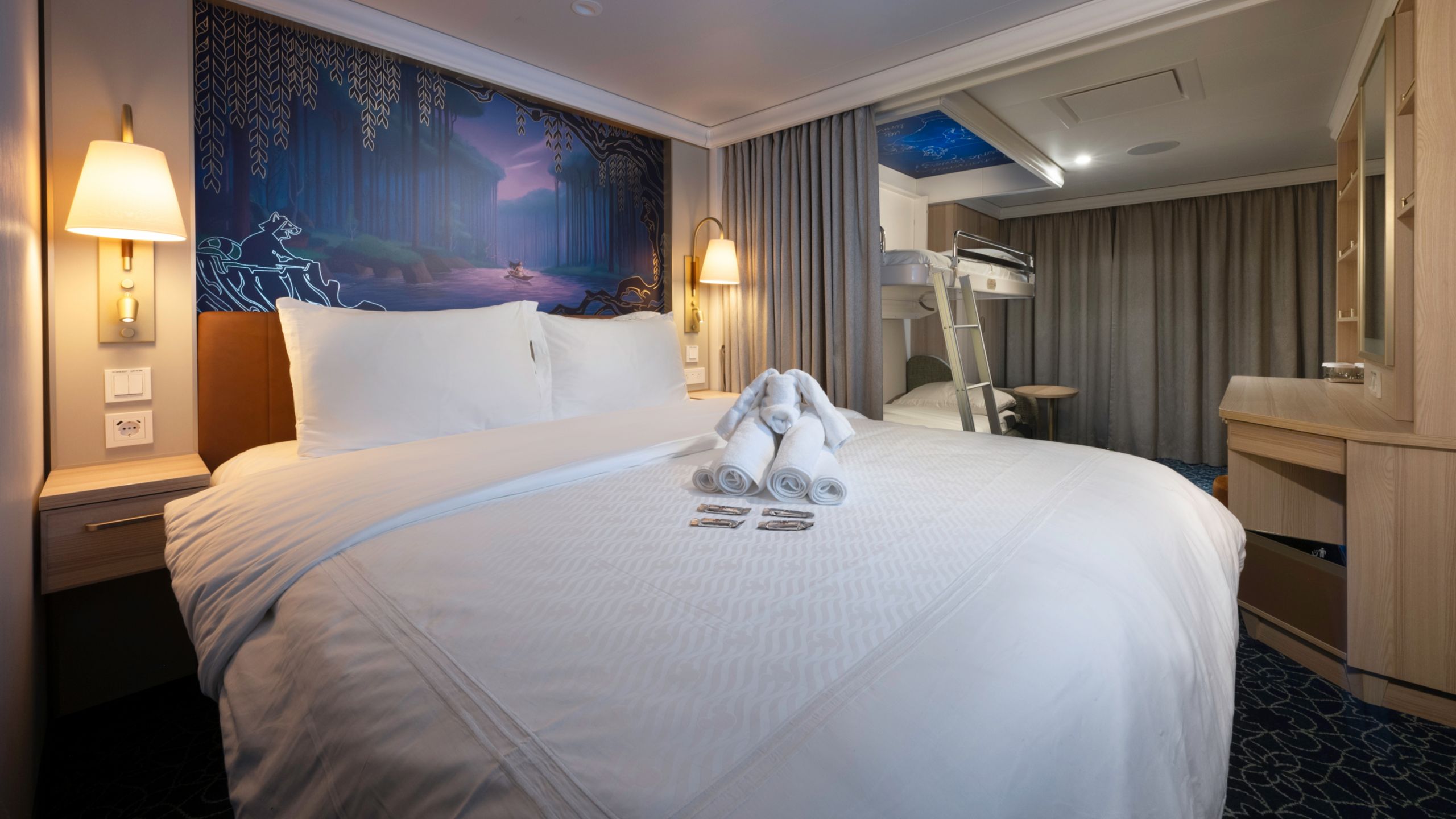 Un camarote del Disney Treasure con escritorio, dos literas, dos mesas de noche, obras de arte inspiradas en Pocahontas y una cama con una toalla doblada con la forma de un elefante