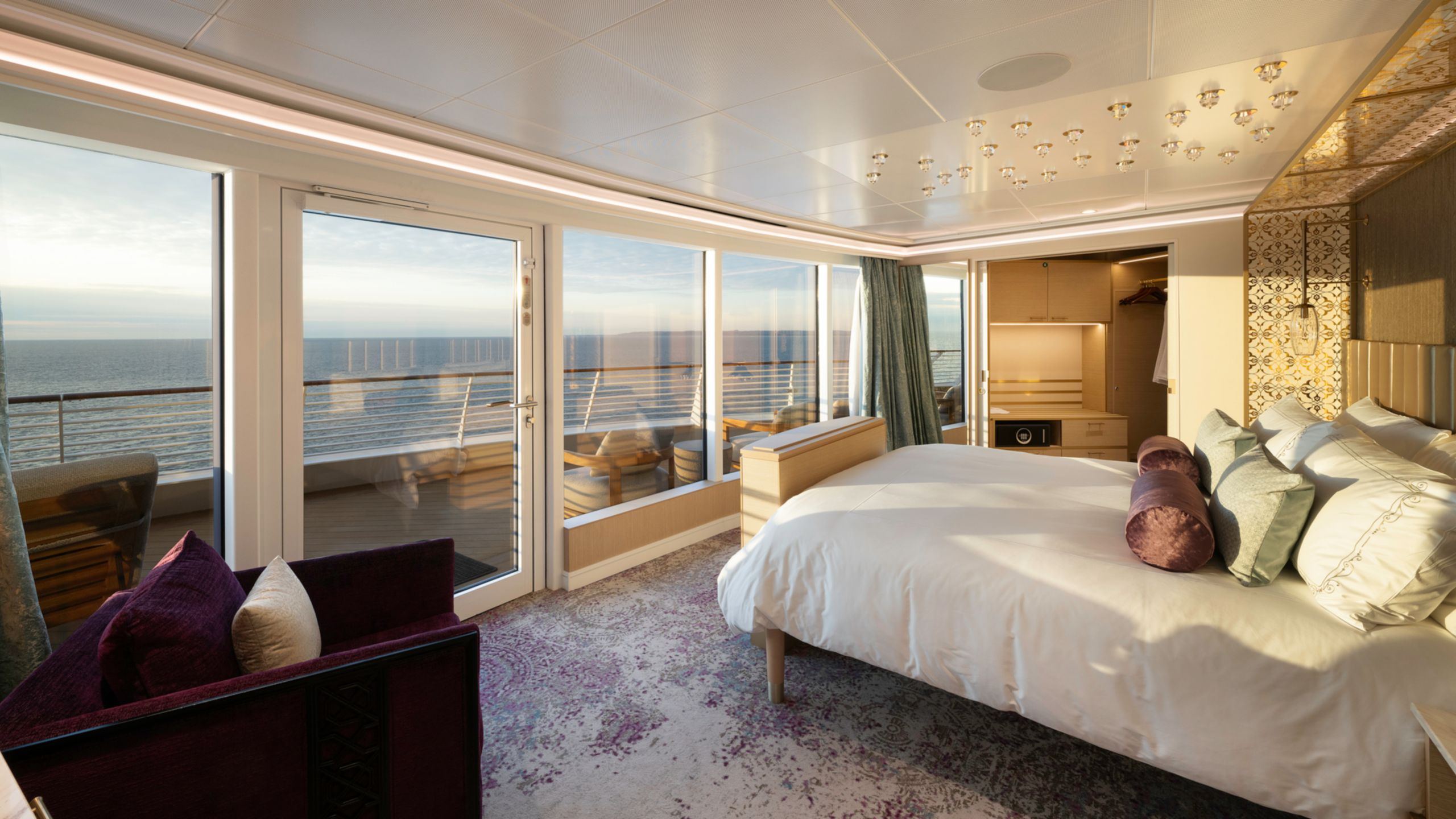 Un camarote del Disney Cruise Line con una cama, armario, escritorio, caja fuerte, silla y un balcón con varias sillas