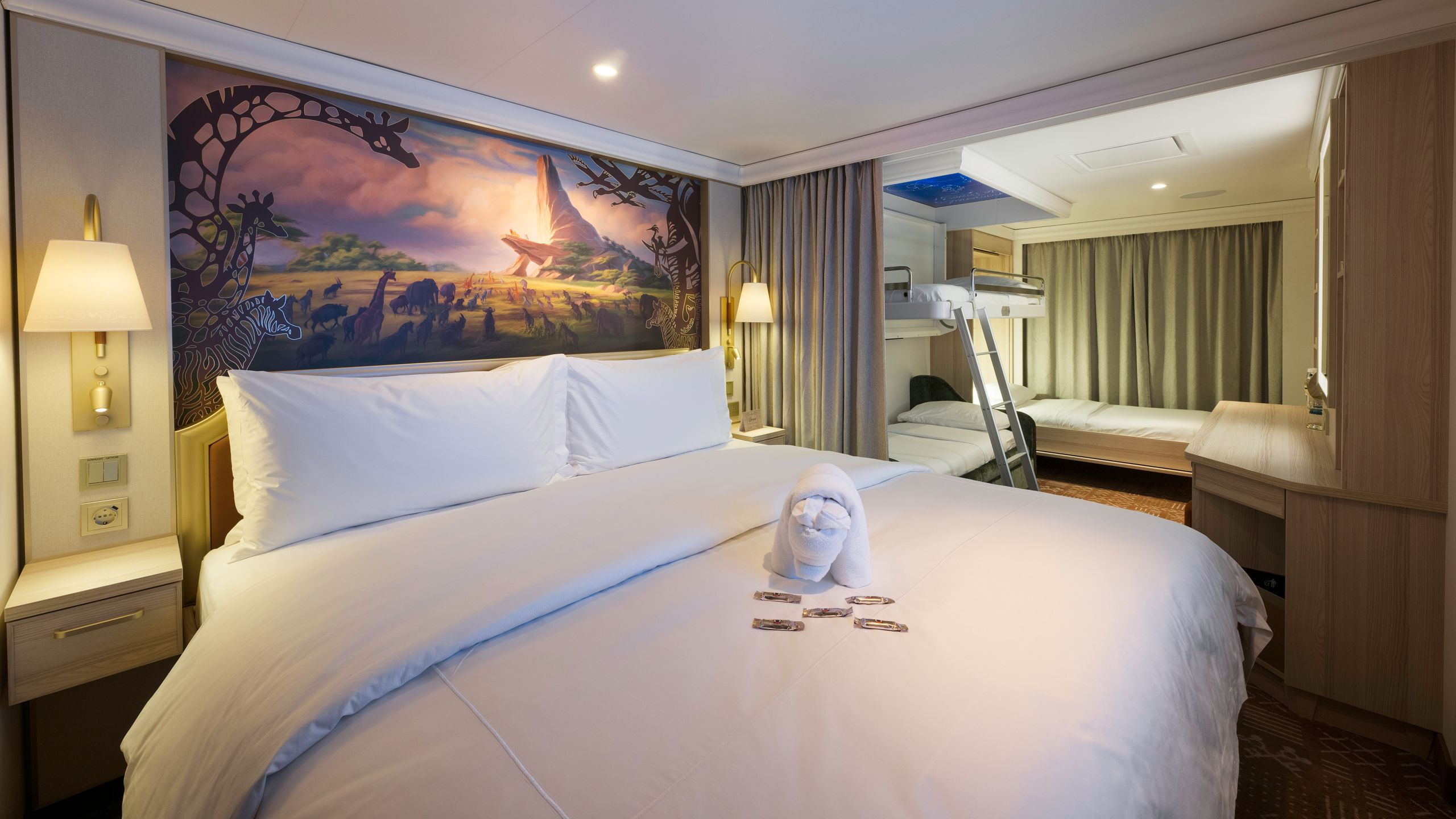 A Disney Cruise Line Verandah Stateroom with a folded towel in the shape of an animal, desk, artwork inspired by The Lion King, 2 beds, 2 nightstands and 2 bunk beds