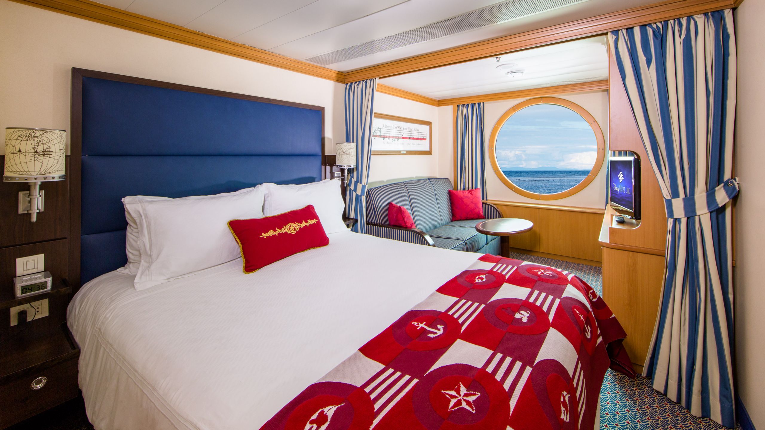 A Disney Cruise Line Deluxe Oceanview Stateroom with a bed, sofa, table, desk, nightstand, TV and a window facing the ocean