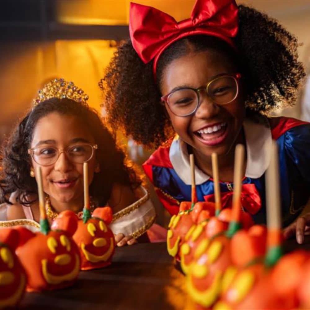Two girls dressed as Snow White and Aurora looking at Halloween themed Mickey Mouse candy apples