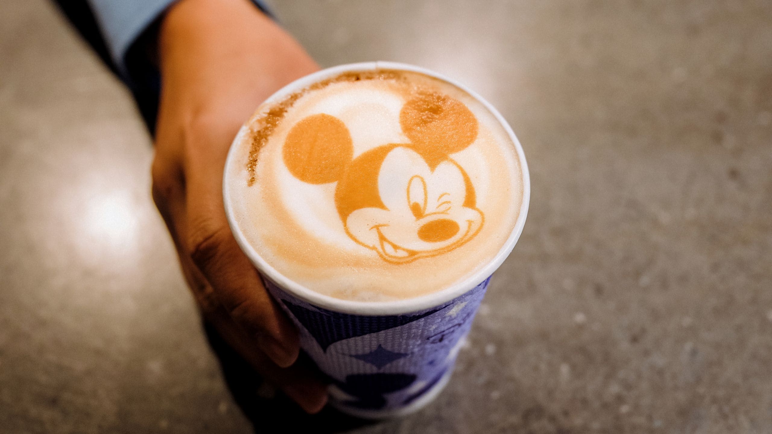 A hand holding a coffee drink with a winking Mickey Mouse drawn into the foam