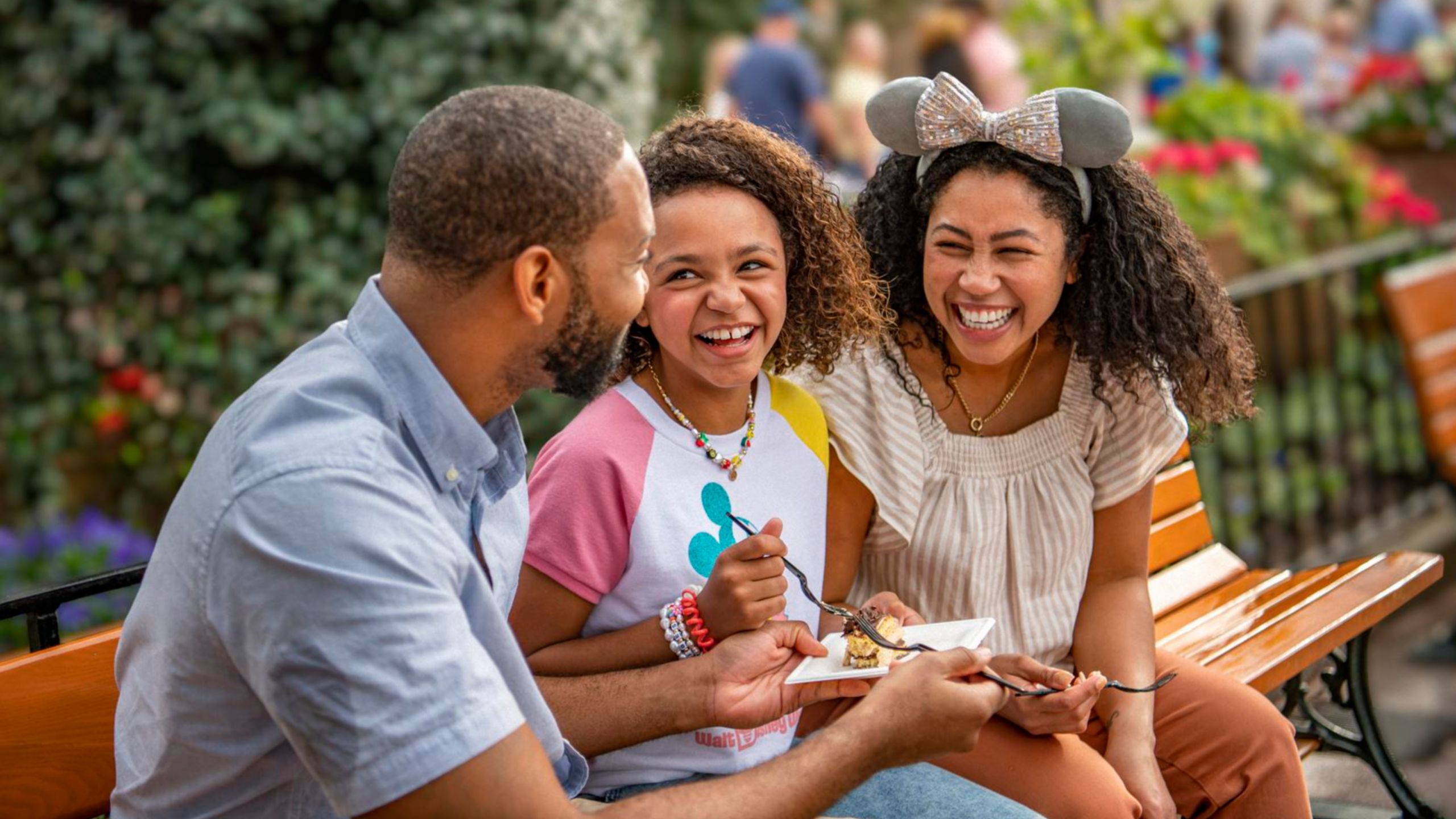 A man, woman and their daughter sitting on a park bench, sharing a snack
