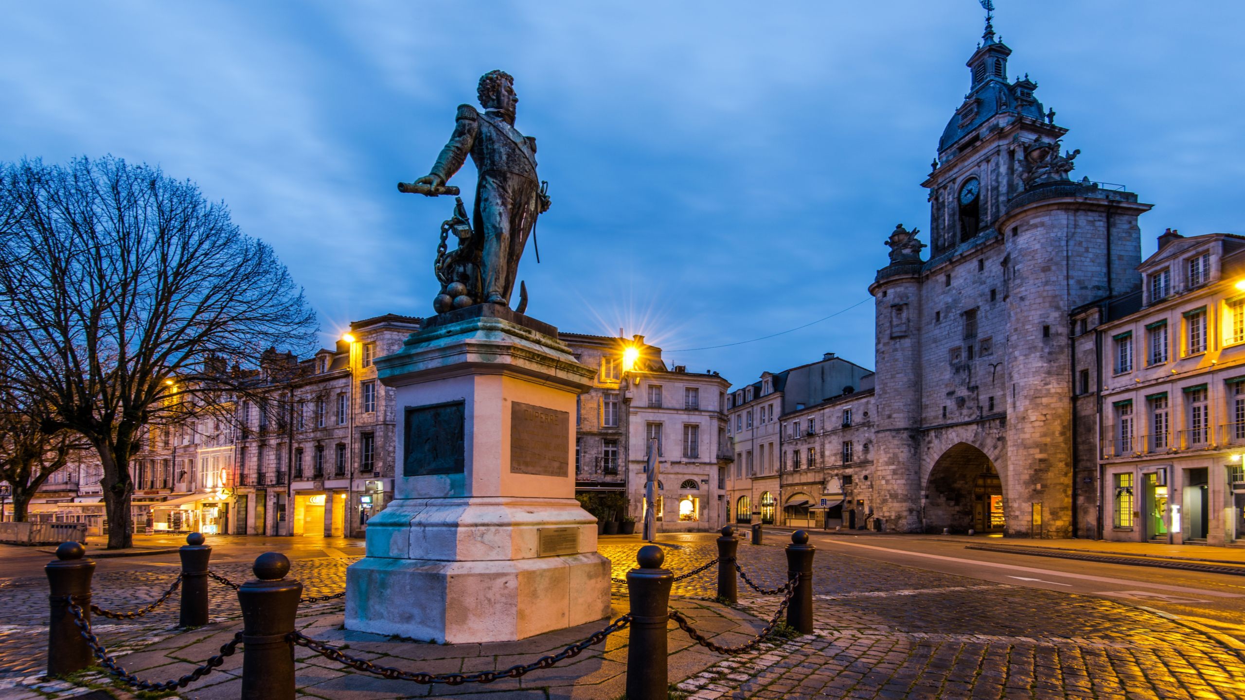 The statue of Admiral Jean Guiton standing in a cobblestone square in La Rochelle, France, near the arched Grosse Horloge stone clock tower