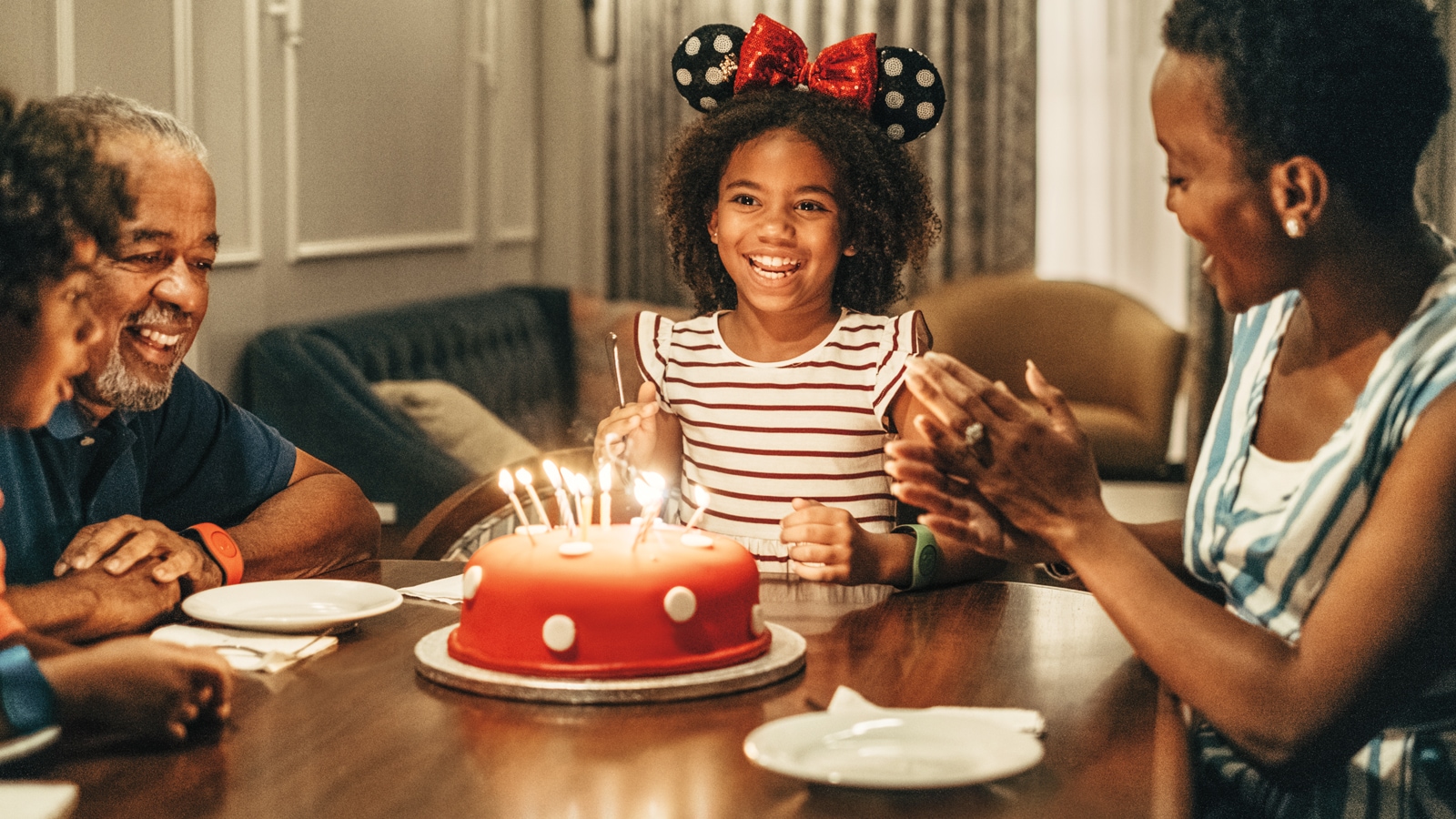 A birthday cake with lit candles on a table in front of a girl wearing a Minnie Mouse ear hat, surrounded by family 