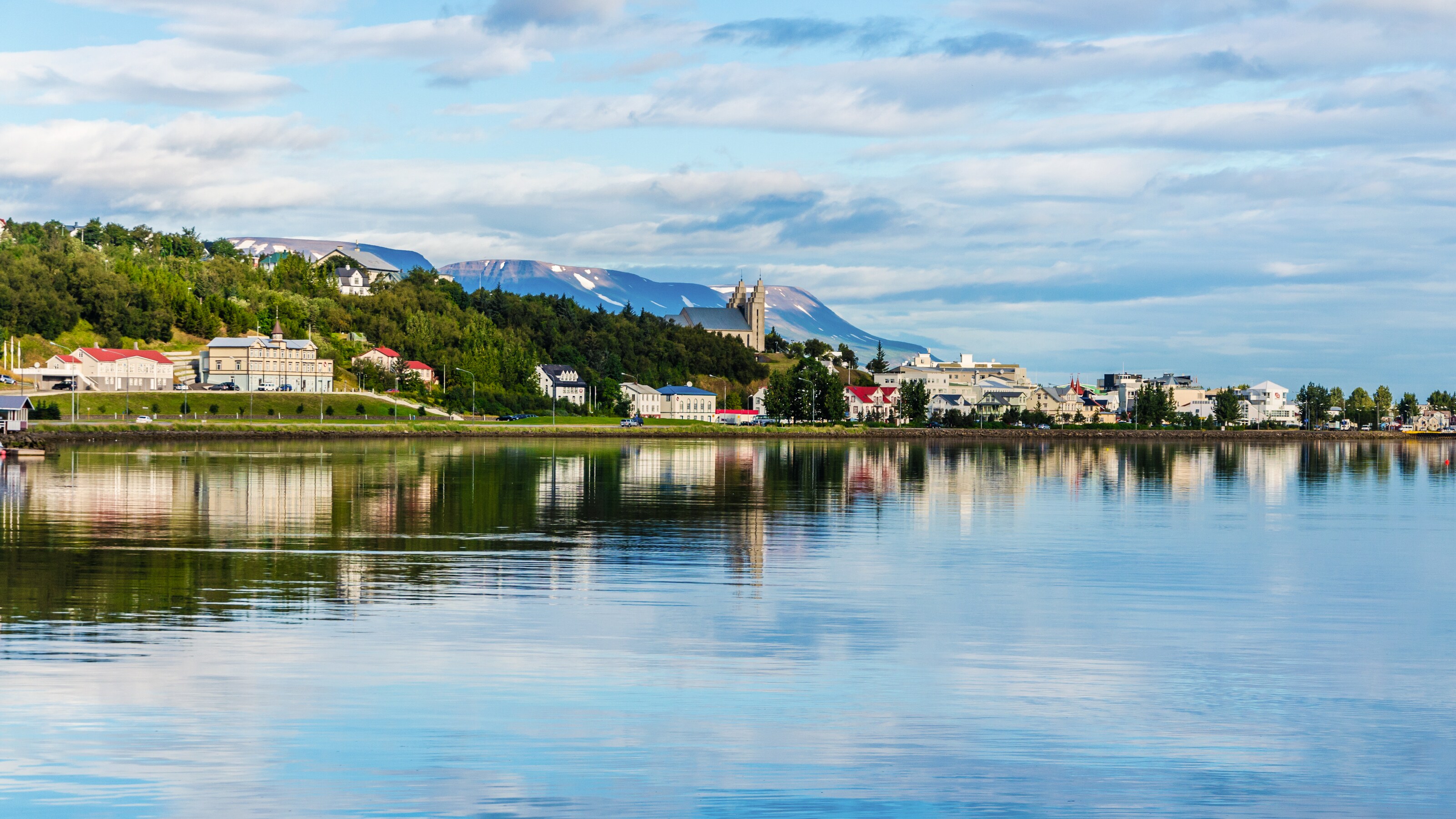 A calm waterfront in Akureyri reflects short buildings, a church and snow capped mountains