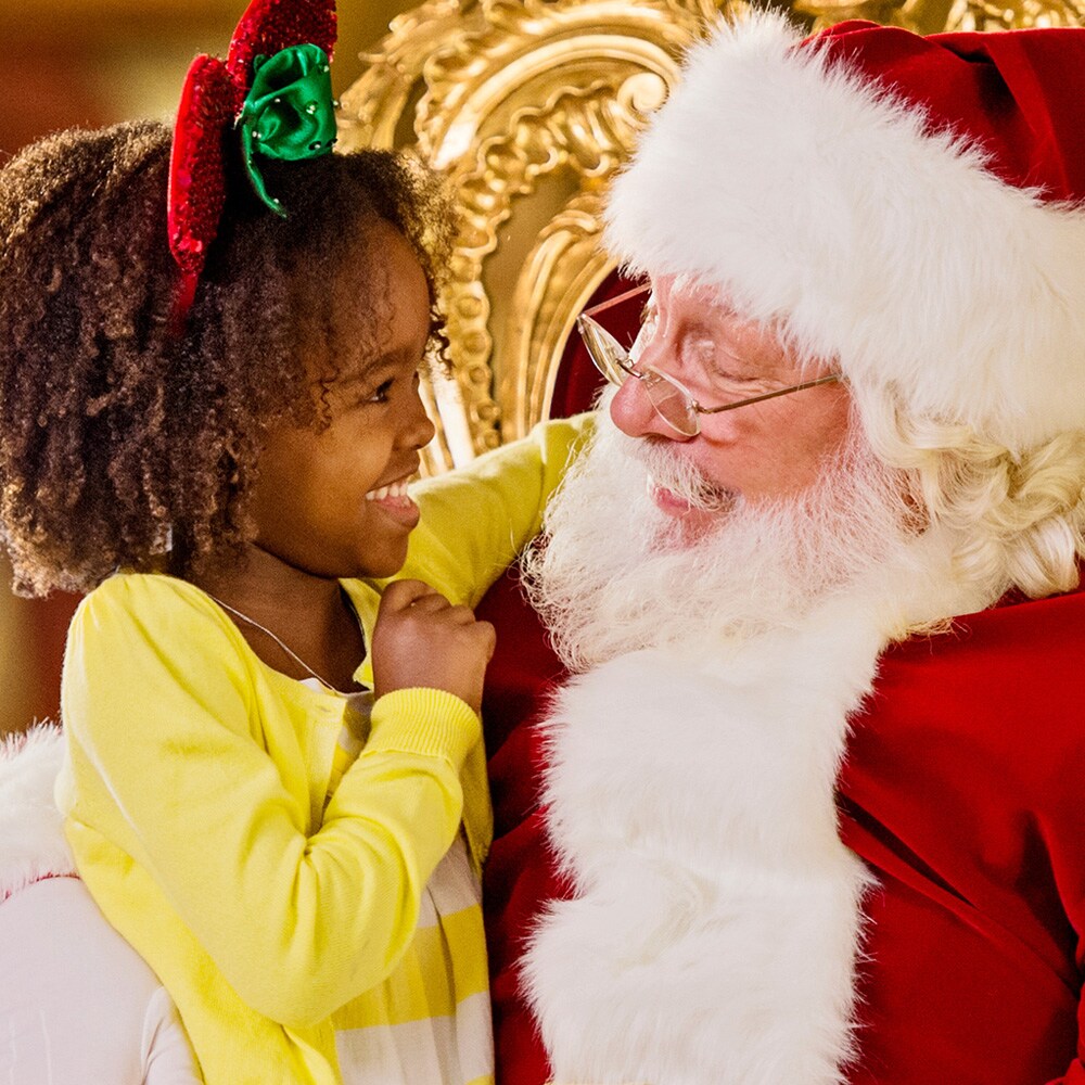 A small girls sits on Santa’s lap and smiles while looking into his eyes